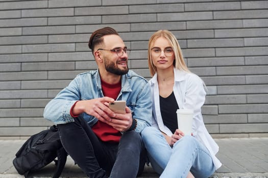 Using phone. Young stylish man with woman in casual clothes sitting outdoors together. Conception of friendship or relationships.