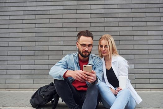 Using phone. Young stylish man with woman in casual clothes sitting outdoors together. Conception of friendship or relationships.