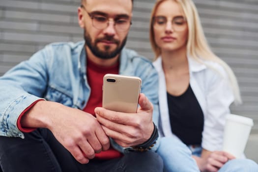 Using phone. Young stylish man with woman in casual clothes sitting outdoors together. Conception of friendship or relationships.