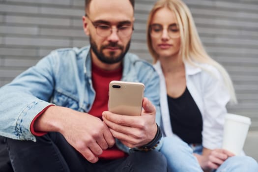 Using phone. Young stylish man with woman in casual clothes sitting outdoors together. Conception of friendship or relationships.