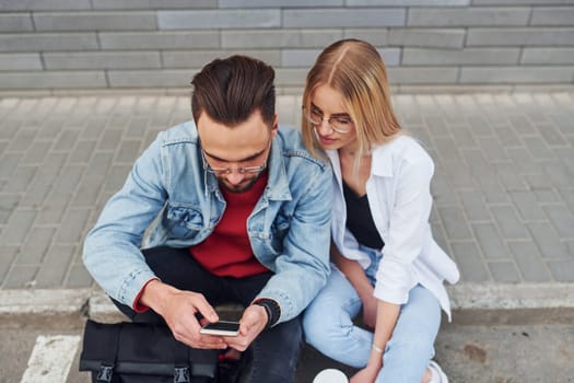 Using phone. Young stylish man with woman in casual clothes sitting outdoors together. Conception of friendship or relationships.