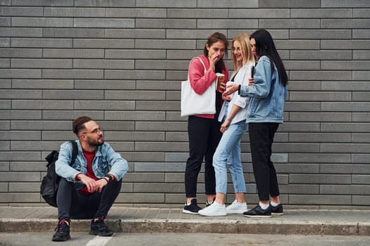 Three women standing and laughing at guy that sitting outdoors near building at daytime.