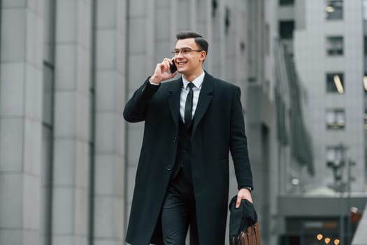 With smartphone. Businessman in black suit and tie is outdoors in the city.