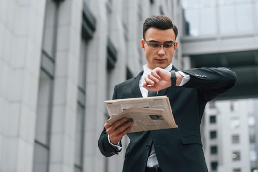 Holding newspaper. Businessman in black suit and tie is outdoors in the city.