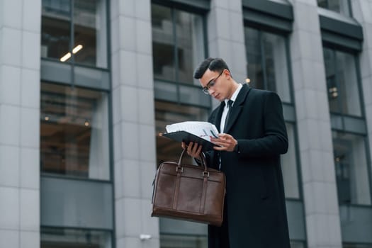 Holding documents. Businessman in black suit and tie is outdoors in the city.