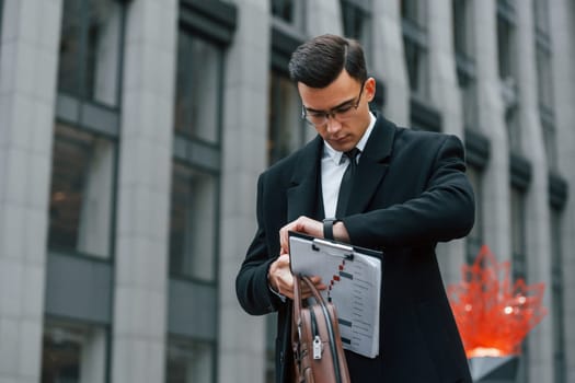 Looking at clock. Businessman in black suit and tie is outdoors in the city.