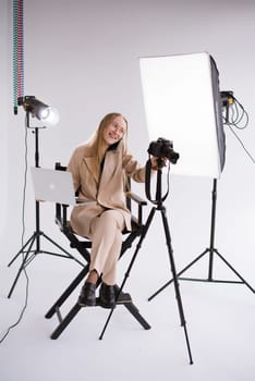 A woman videographer, a blonde with a camera and laptop apple, happy talking on the phone and smiling in the studio. Wearing a formal nude pantsuit on a white isolated background