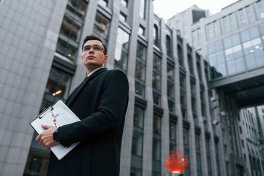 With notepad in hands. Businessman in black suit and tie is outdoors in the city.