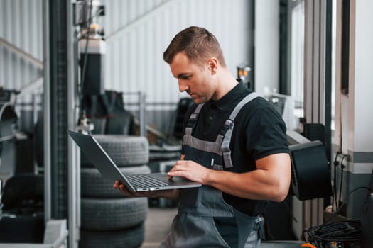 Holding laptop in hands. Man in uniform is working in the auto service.