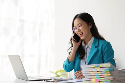 Asian Woman entrepreneur busy with her work in the office. Young Asian woman talking over smartphone or cellphone while working on computer at her desk...