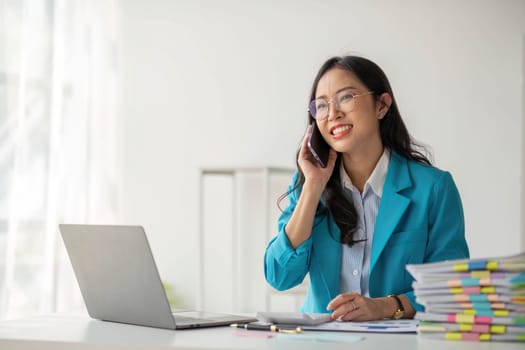 Asian Woman entrepreneur busy with her work in the office. Young Asian woman talking over smartphone or cellphone while working on computer at her desk...