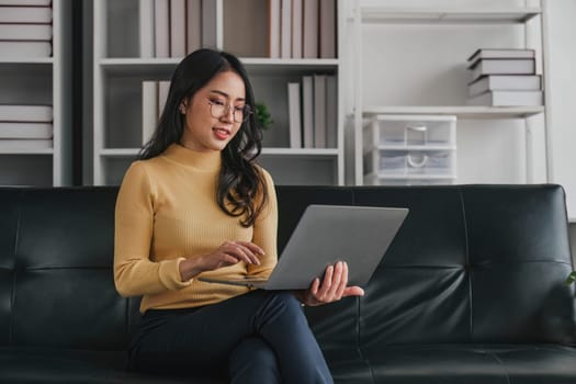 Young asian woman in good spirits working on laptop at home while sitting on the couch..