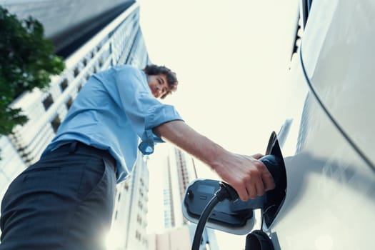 Fisheye view focus on hand insert EV charger plug into electric car with blur background of progressive modern city lifestyle-businessman recharge his EV car with residential building condo background