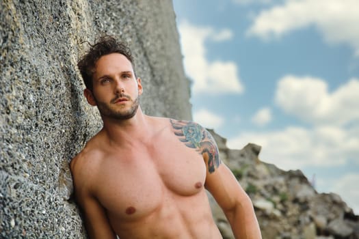 Attractive muscular shirtless athletic man standing on rock next to water by sea or ocean shore, looking at camera in a cloudy summer day