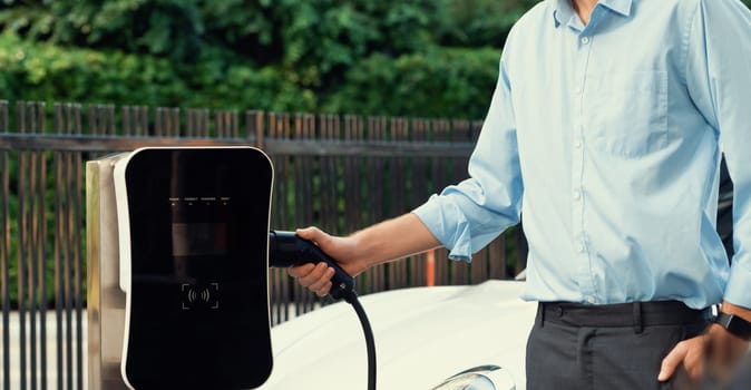 Closeup progressive man holding EV charger plug from public charging station for electric vehicle with background of residential building as concept eco-friendly sustainability energy car concept.