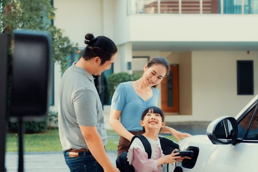 Progressive young parents and daughter with electric vehicle and home charging station. Green and clean energy from electric vehicles for healthy environment. Eco power from renewable source at home.