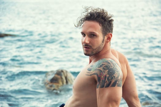Attractive muscular shirtless athletic man sitting next to water by sea or ocean shore, looking at camera in a cloudy summer day