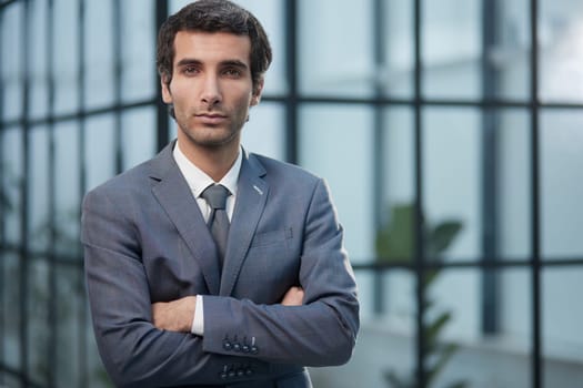 a dark-haired man in an office in a suit stands with crossed arms in front of him