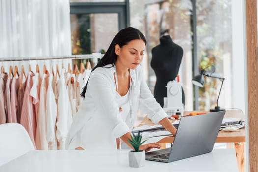 Using laptop. Seamstress is in her office that is with different clothes.