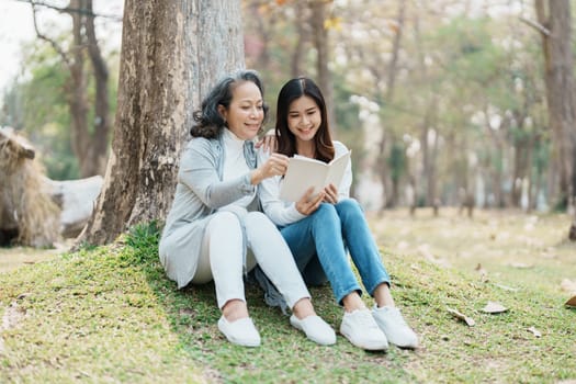 Grown daughter with aging mother expressing love and looking at memory notebook under tree in public park.