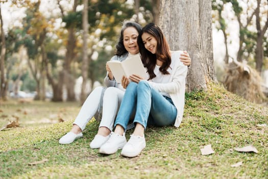 Grown daughter with aging mother expressing love and looking at memory notebook under tree in public park.