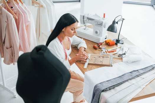Sitting by the table. Seamstress is in her office that is with different clothes.
