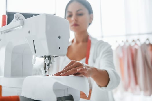 Working by using sewing machine. Seamstress is in her office that is with different clothes.
