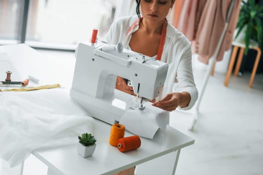 Working by using sewing machine. Seamstress is in her office that is with different clothes.