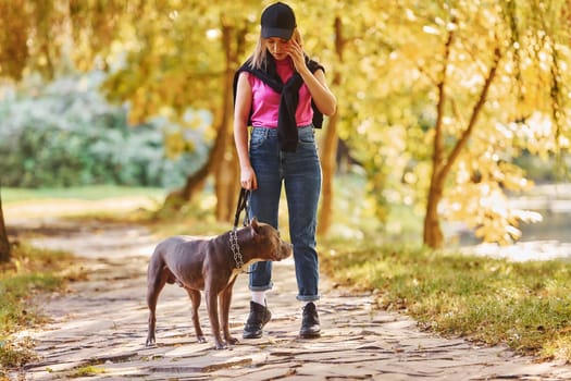 Standing together. Woman in casual clothes is with pit bull outdoors.