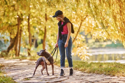 Standing together. Woman in casual clothes is with pit bull outdoors.