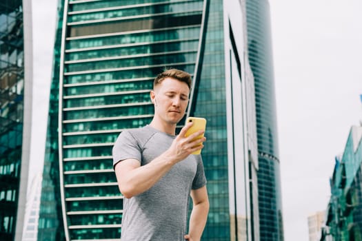 Tired Young man runner standing and using mobile phone and relaxing after sport training. Holding water bottle while doing fitness workout in city urban street, cloudy sky at summer