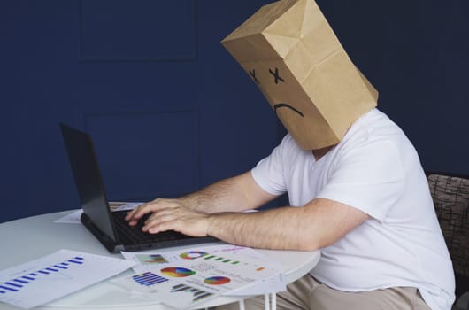 A male businessman in a white shirt with a paper bag on his head, with a sad smiley face drawn, works at a computer, with documents. Emotions and gestures.