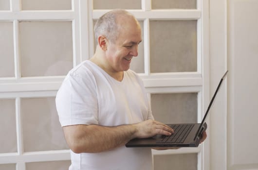 A man holds a laptop in his hands, stands at home and has a lively conversation on a video call.