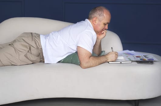 A businessman lies on a sofa and looks at data trends on charts and graphs, makes notes in a notebook. Distant work