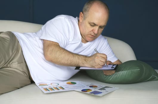 A businessman lies on a sofa and looks at data trends on charts and graphs in a tablet, makes notes in a notebook. Distant work