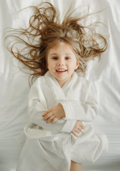 Joyful girl in a white bathrobe lies in bed, looks at the camera. View from above.