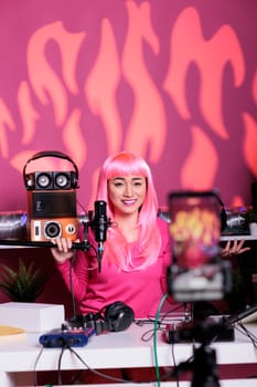 Asian woman with pink hair talking with subscribers using professional microphone while filming broadcast in vlogging studio. Influencer recording podcast with mobile phone camera, live streaming