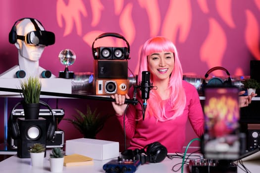 Cheerful woman recording podcast using professional streaming equipment while standing in broadcast studio, posting vlog on social media to gain fans. Asian creator filming vlog with phone camera