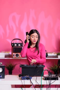 Asian artist with headphones having fun while playing techno music using professional mixer console, standing at dj table in studio with pink background. Musician performing electronic song in club