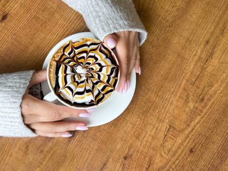 Coffee with spider web pattern on top of foam in white cup and girl's hands on wooden table background, top view