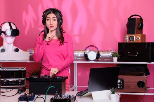 Performer putting forefinger on lips gesturing secrecy sign while performing electronic music in studio over pink background. Musical artist playing sounds to produce melody at mixer console