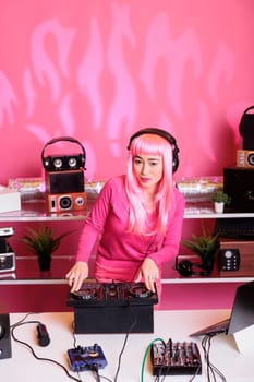 Artist with pink hair wearing headset while playing electronic music using professional mixer console, enjoying performing in night club. Asian musician standing at dj table mixing sounds