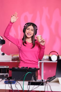 Happy performer playing techno sound at professional mixer console while drinking coffee, enjoying playing songs at night in club. Asian musician creating musical performance with remix music