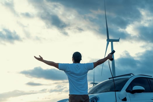 Progressive man spread arm with electric car recharge energy from charging station on green field powered by wind turbine concept of future sustainable energy.