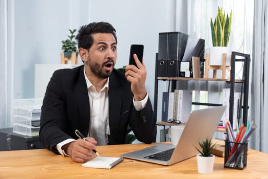 Diligent businessman busy talking on the phone call with clients while working with laptop in his office desk as concept of modern hardworking office worker lifestyle with mobile phone. Fervent