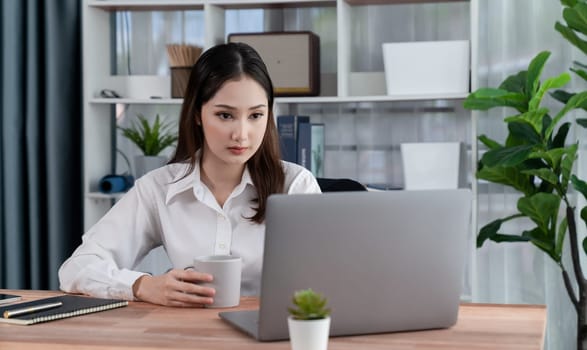 Young asian enthusiastic businesswoman at modern office desk using laptop to work with a cup of coffee. Diligent and attractive office lady working on computer notebook in her office work space.