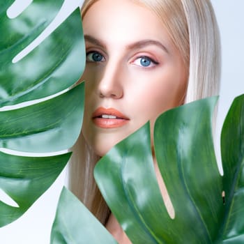 Closeup facial portrait personable woman with perfect smooth makeup holding green monstera leaves and cover her face as natural healthy skincare treatment. Tropical nature and beauty concept