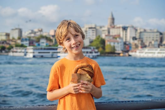 Boy in Istanbul having breakfast with Simit and a glass of Turkish tea. Glass of Turkish tea and bagel Simit against golden horn bay and the Galata Tower in Istanbul, Turkey. Turkiye. Traveling with kids concept.