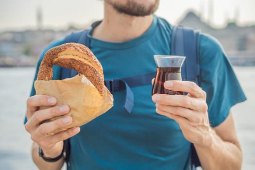 Man in Istanbul having breakfast with Simit and a glass of Turkish tea. Glass of Turkish tea and bagel Simit against golden horn bay in Istanbul, Turkey. Turkiye.