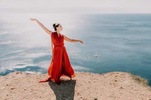 Side view a Young beautiful sensual woman in a red long dress posing on a rock high above the sea during sunrise. Girl on the nature on blue sky background. Fashion photo.